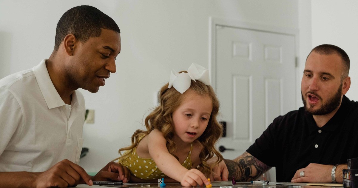 A young girl and two adults leaning over a colorful board game at a kitchen table