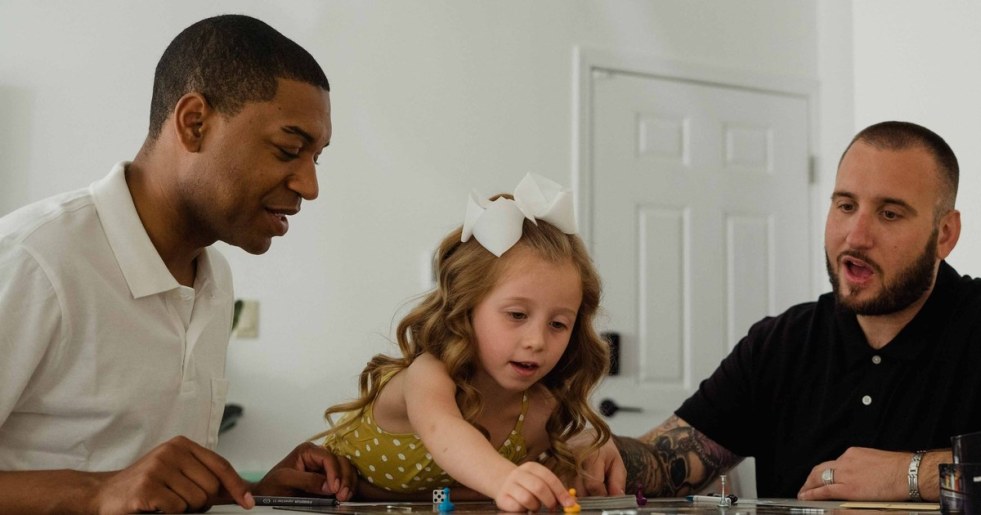 A young girl and two adults leaning over a colorful board game at a kitchen table