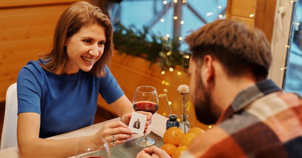 A young couple smiling over wine and a card game by string lights