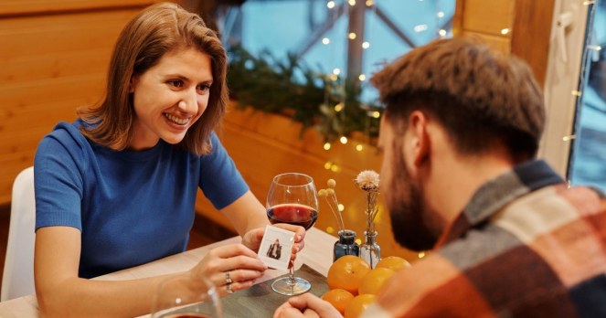A young couple smiling over wine and a card game by string lights