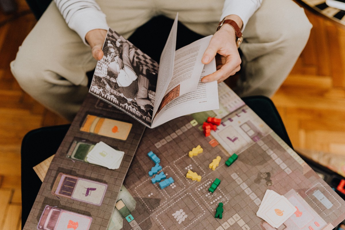 A man in a cream sweater reads a game rulebook above an open board with colorful wooden pieces