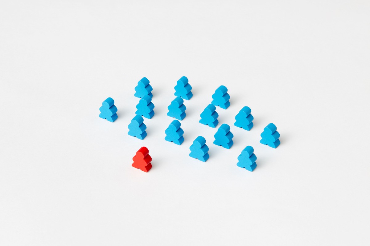 One red wooden meeple standing out among a group of blue wooden meeples on a white surface