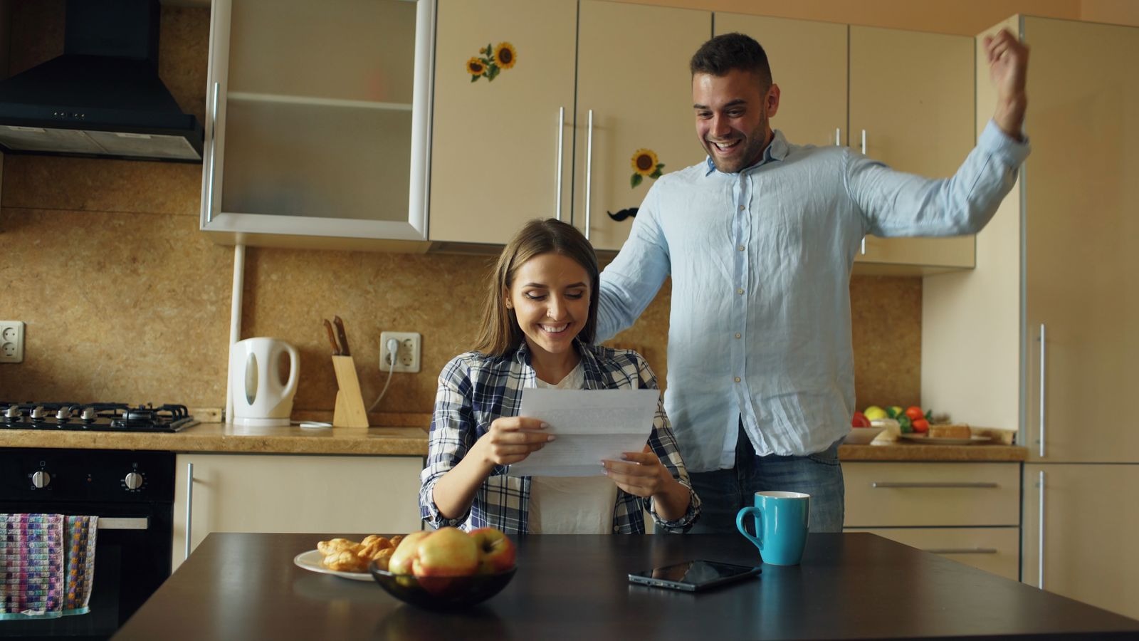 A woman reading a letter in a kitchen while a man behind her cheers