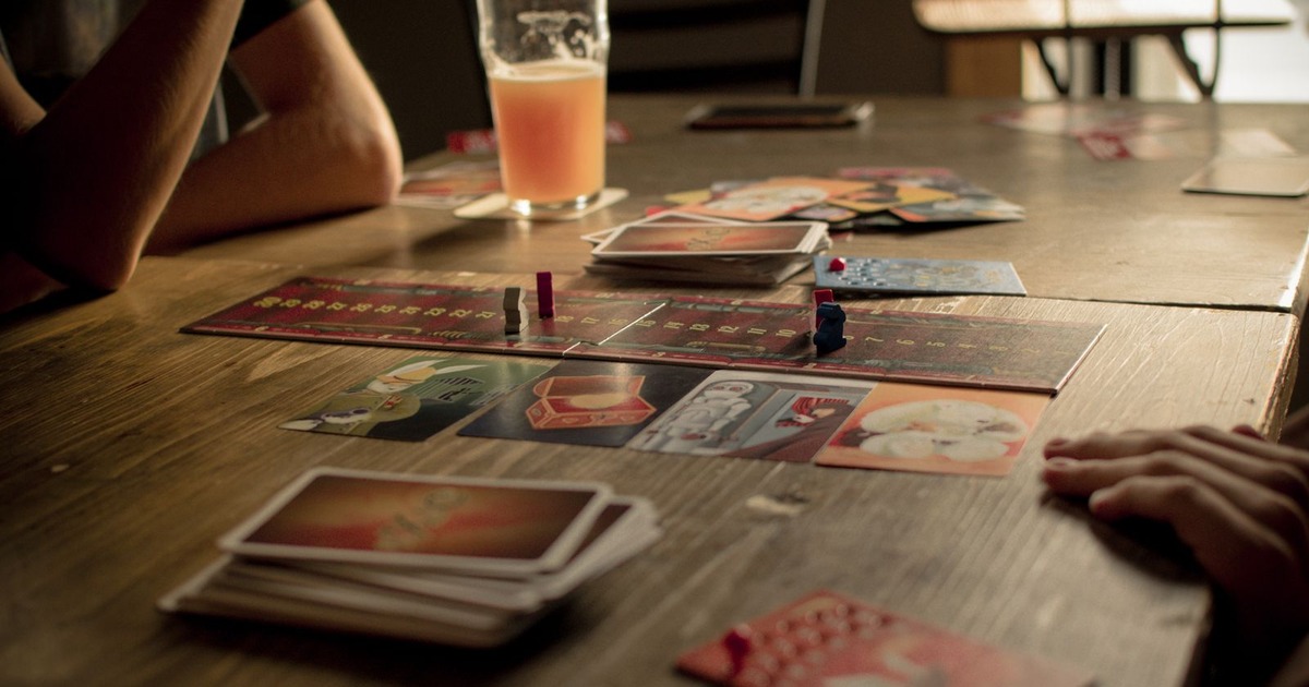 A board game in progress on a wooden table with cards, a drink and a player's hand at the edge