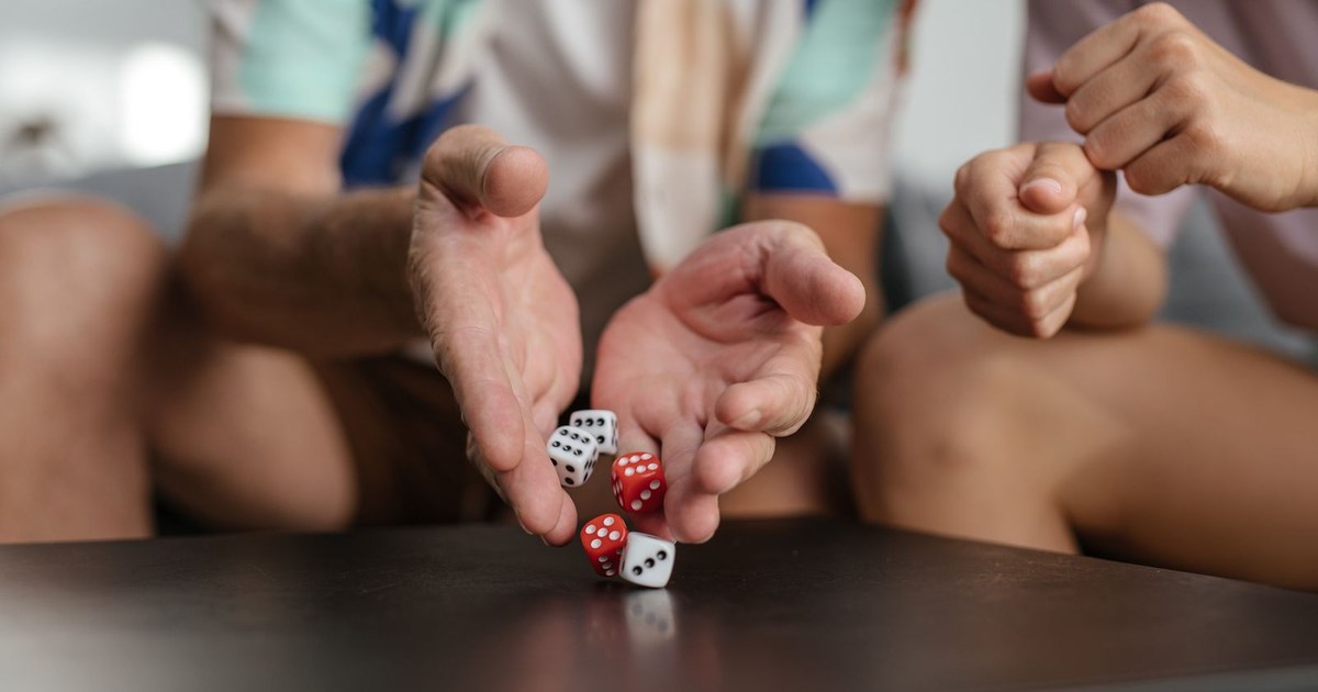 Hands tossing red and white dice onto a dark coffee table while friends watch