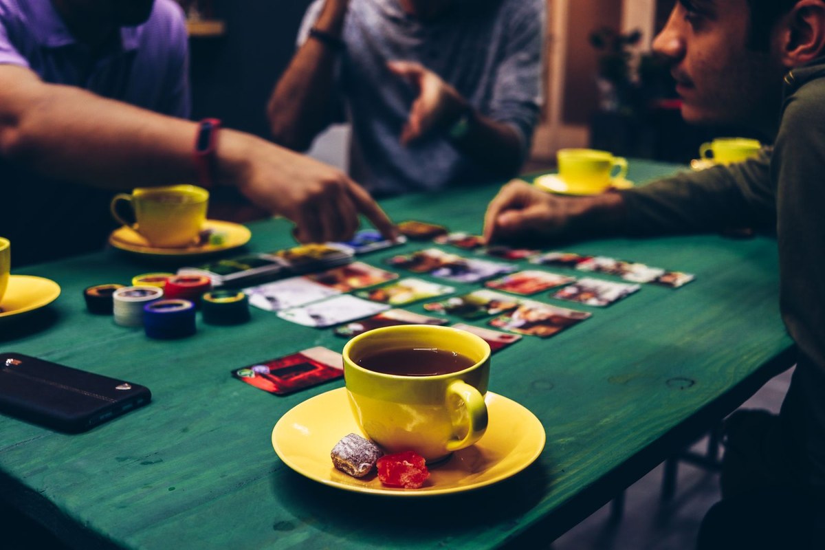 Friends reach across a green table of picture cards and chips beside a yellow cup of tea