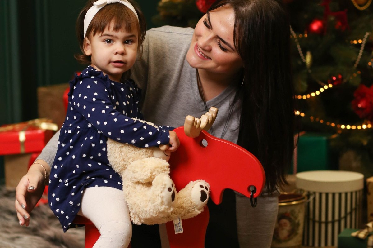 A mother holding a toddler on a red wooden moose rocker in front of a Christmas tree with wrapped gifts